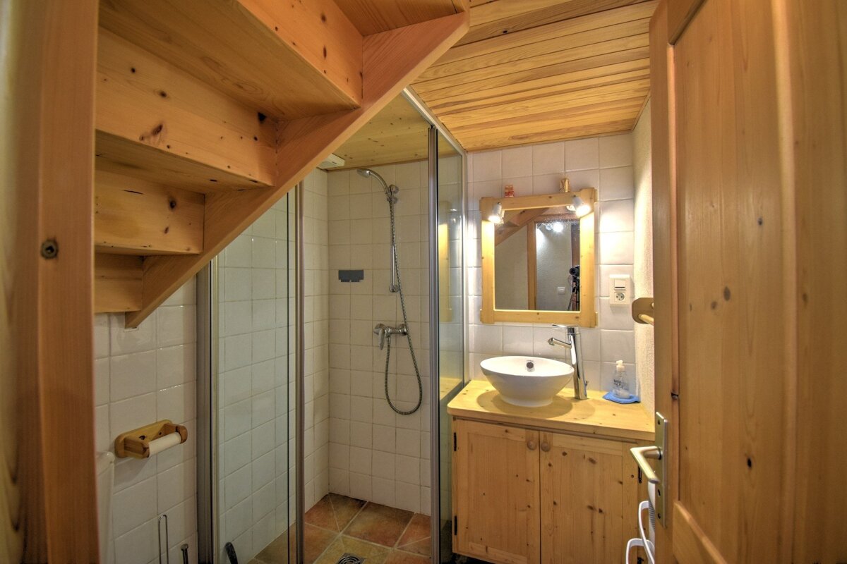A rustic bathroom with light wood stairs overhead, a shower stall, a round basin on a wooden vanity, and a wooden framed mirror.