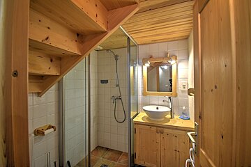 A compact bathroom with a shower, wooden vanity & vessel sink, beneath an overhead wooden staircase and paneled ceiling.