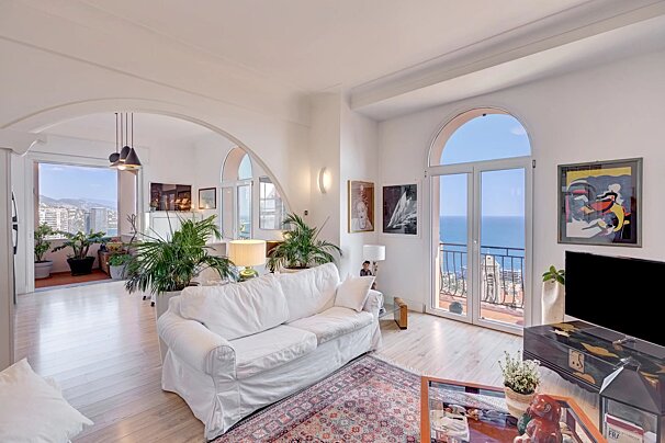 Bright, airy living room with a white sofa, houseplants, and arched doors leading to a balcony overlooking the sea.