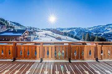 Wooden balcony with carved tree details overlooking a sun-drenched ski resort and snowy mountains.