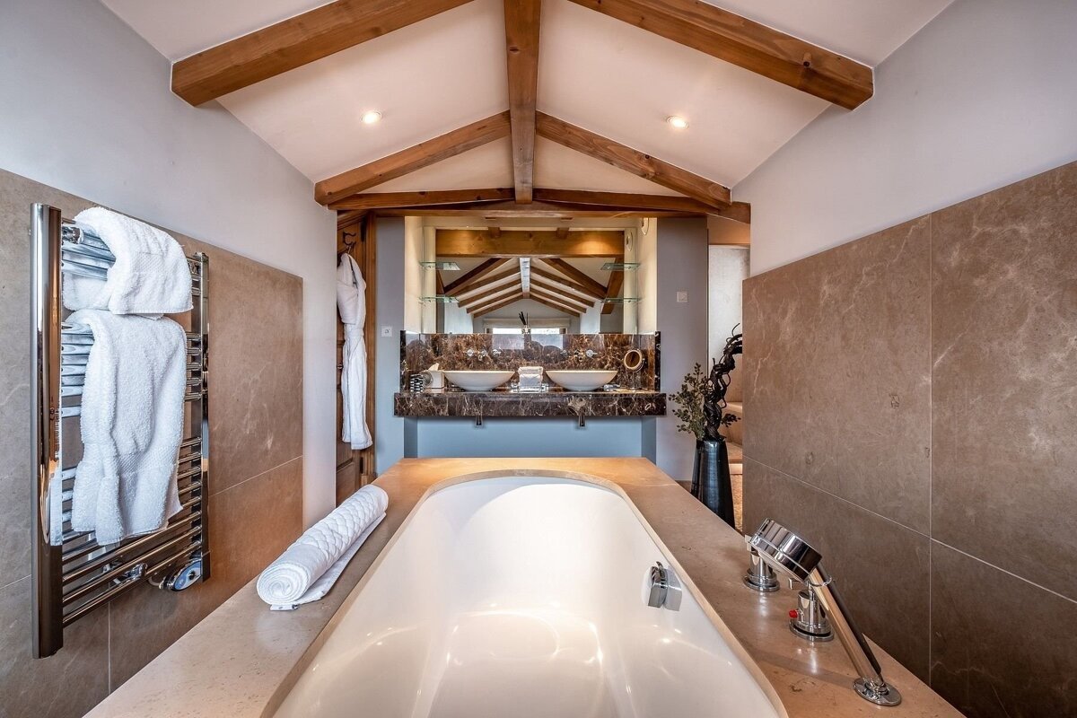 An upscale bathroom featuring a large built-in tub, dual vessel sinks with a marble countertop, and vaulted ceiling with exposed wooden beams.