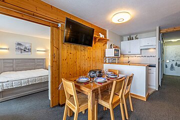 A cozy, open-plan ski lodge apartment featuring a wooden dining table, kitchenette, and a bedroom visible through a wooden archway, with mountain decor.