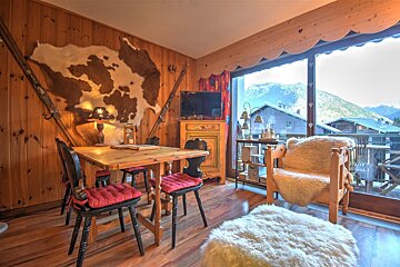 Cozy wooden chalet interior with a dining table, rustic decor, sheepskin seating, and mountain views outside.