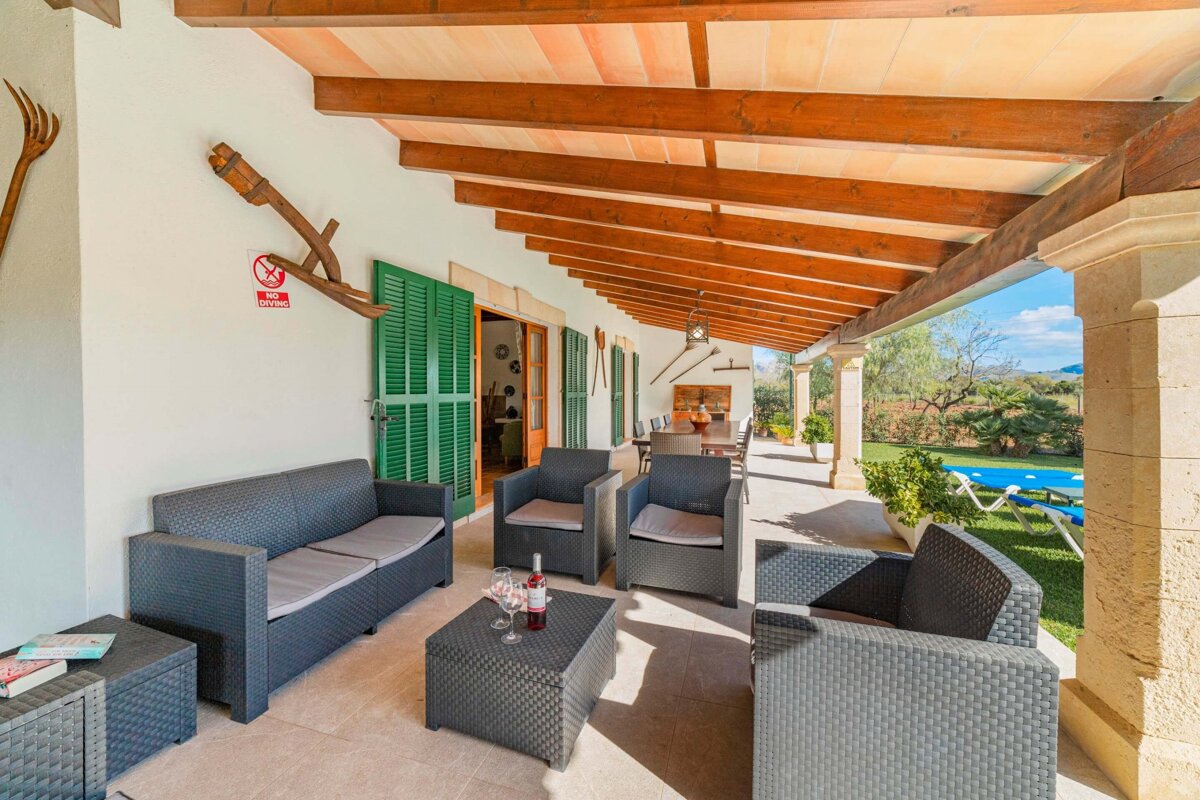 A spacious covered patio with dark wicker furniture, exposed wooden beams, and green shutters, overlooking a vibrant garden and swimming pool.