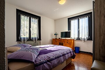 Bright bedroom featuring a bed with purple bedding, two black-framed windows, and a wooden dresser with a TV.