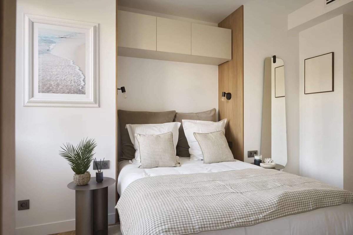 A cozy, modern bedroom with a built-in bed niche, neutral gingham bedding, overhead cabinets, wooden accents, a beach art print, and a minimalist mirror.