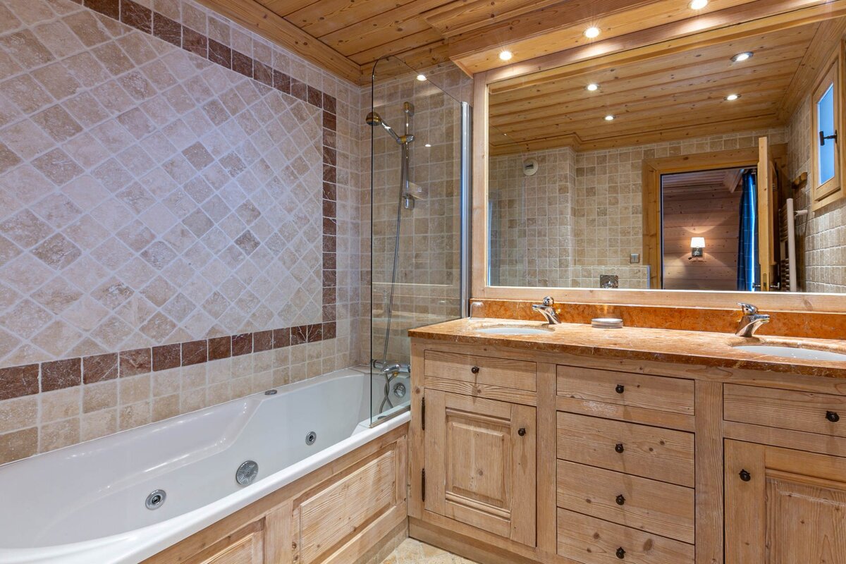 A warm, rustic bathroom features a wood-paneled ceiling, tiled walls, a bathtub with a shower, and a wooden vanity with double sinks.