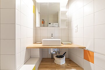 Bright, modern, white-tiled bathroom with a wooden counter and white vessel sink. A mirror cabinet above reflects a washing machine; yellow/orange accents.