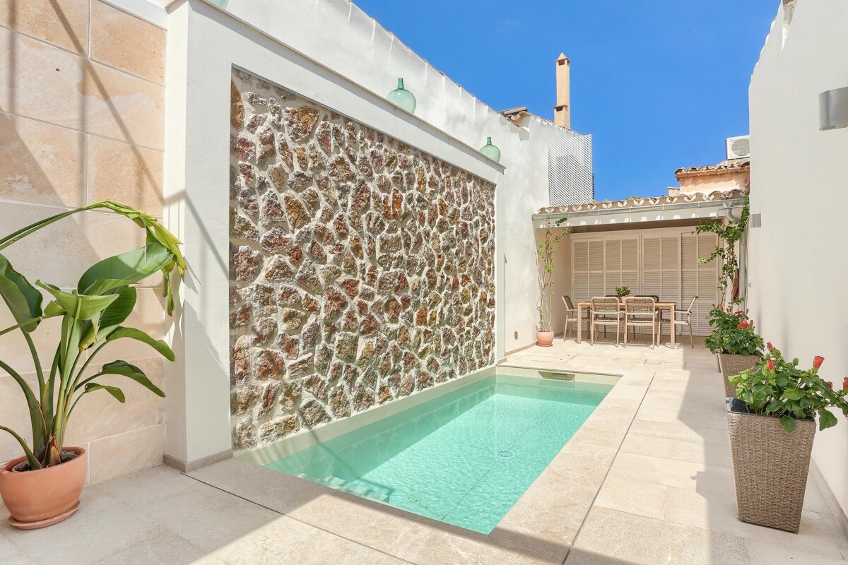 A tranquil outdoor space featuring a small pool by a rustic stone wall, with an outdoor dining area under a trellis, all under a bright blue sky.