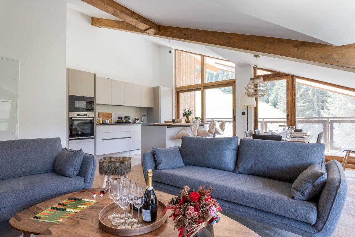 Modern open-plan living room with grey sofas, kitchen, dining area, and large windows overlooking a forest, featuring exposed wooden beams.