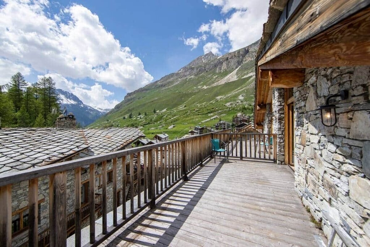 View from a wooden chalet balcony overlooking a stone-roofed alpine village and lush green mountains under a cloudy blue sky.