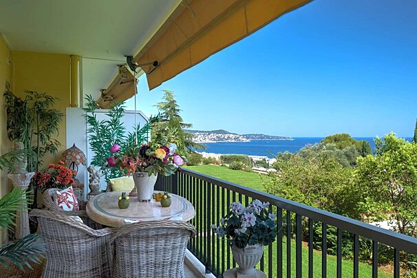 A sunny balcony adorned with wicker furniture and vibrant flowers, boasting a stunning panoramic view of a sparkling blue sea and green hills.