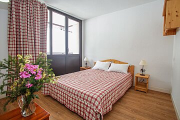A cozy bedroom with a double bed, red and white plaid bedding, wooden furniture, a large window with matching plaid curtains, and a vase of flowers.
