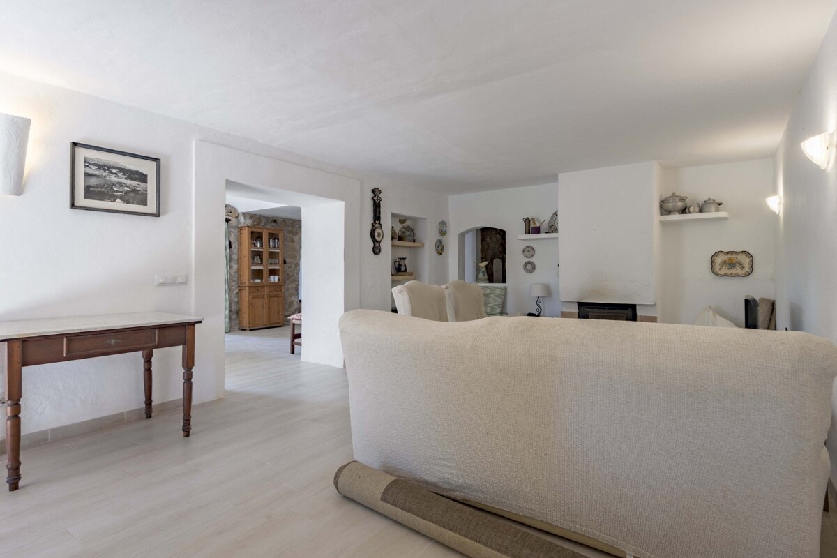 A spacious, bright living room with white walls, a light floor, and a cream sofa in the foreground, leading to other rustic-style rooms.