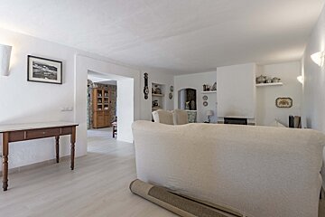 A spacious, bright living room with white walls, a light floor, and a cream sofa in the foreground, leading to other rustic-style rooms.