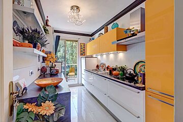 A vibrant, modern kitchen with glossy orange upper cabinets, white lower units, shiny tiled floor, and a balcony view to greenery.