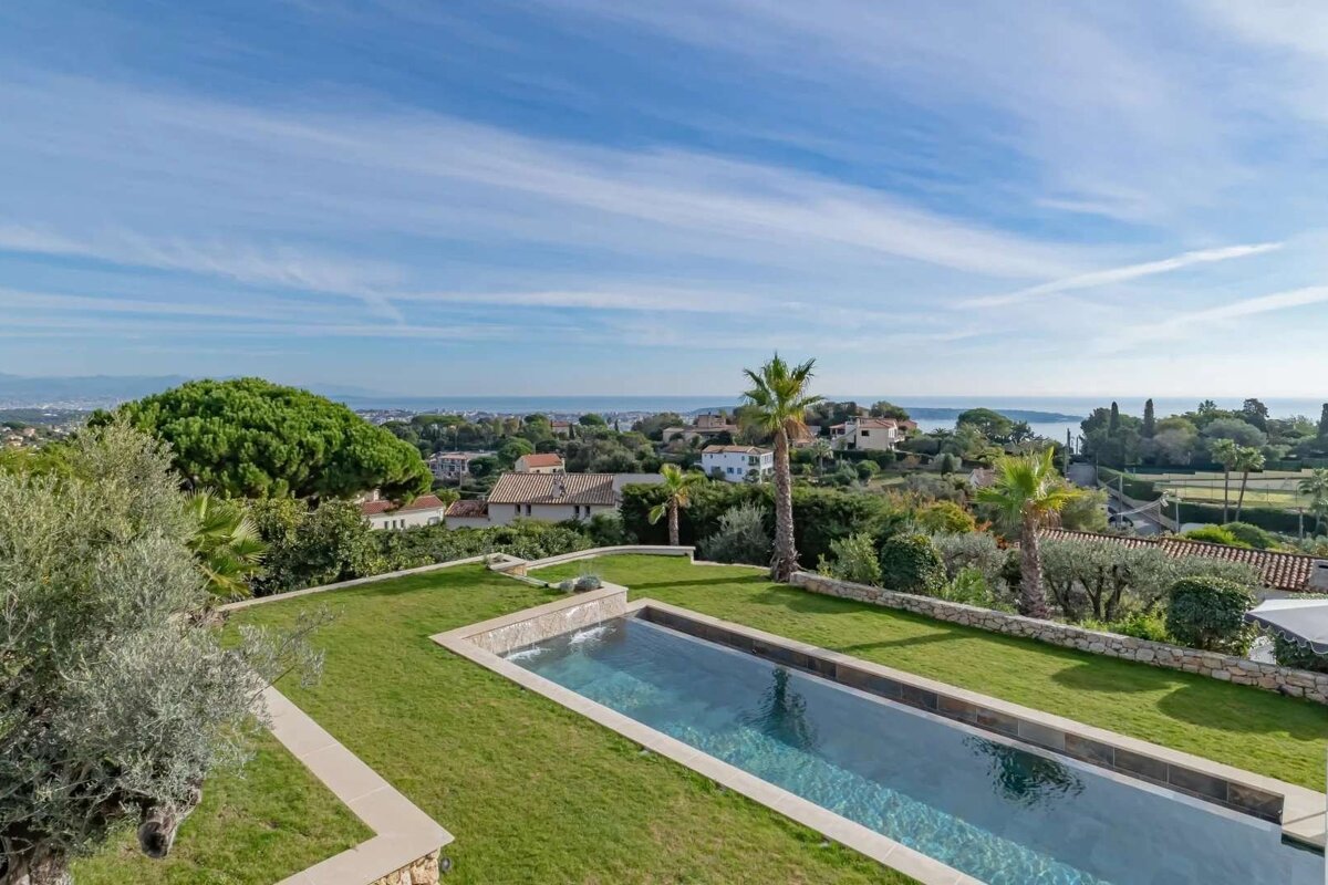 Infinity pool on a green lawn, overlooking a coastal town and sea under a blue sky.