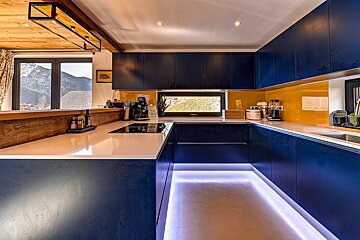 A stylish modern kitchen featuring blue cabinetry, a vibrant yellow backsplash, white countertops, and glowing purple under-cabinet lights. A mountain view is visible from the window.