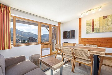 Bright, modern living/dining room with panoramic mountain views through large windows. Features wooden furniture, sofa, and TV.