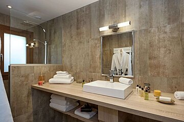 A modern bathroom with earthy-toned tiles, a wooden vanity, and a rectangular white sink. White towels and toiletries are neatly arranged. A mirror reflects a white robe.