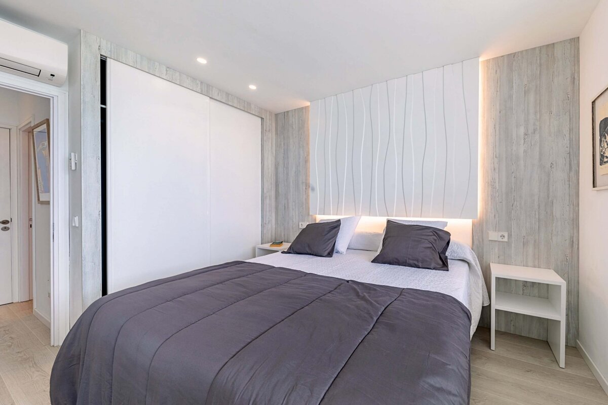 A modern bedroom features a large bed with dark grey bedding, a backlit white wavy headboard, built-in closet, and light wood accent walls.