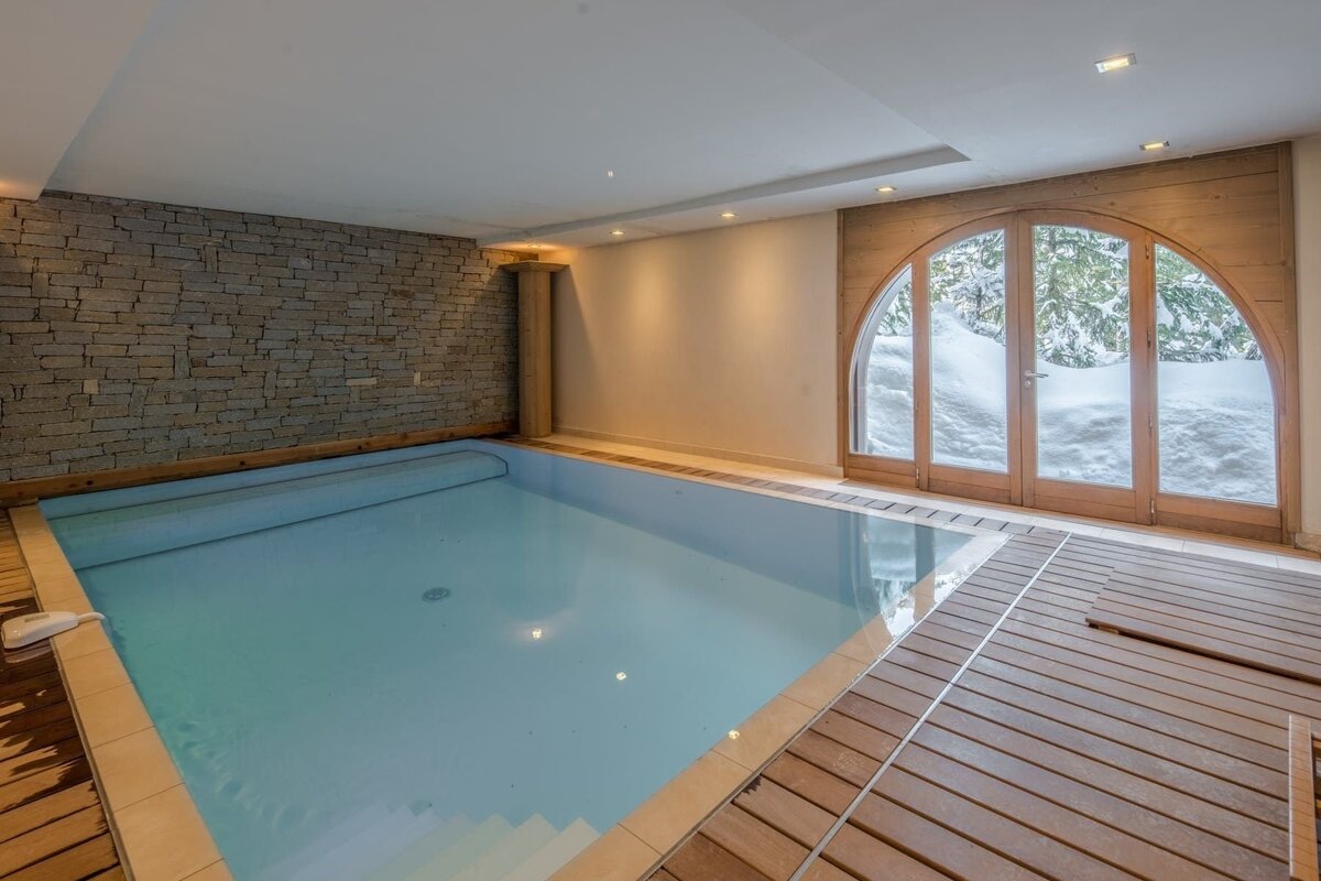 A warm indoor pool with a stone accent wall and wood decking. Large arched windows offer a serene view of a snowy winter landscape outside.