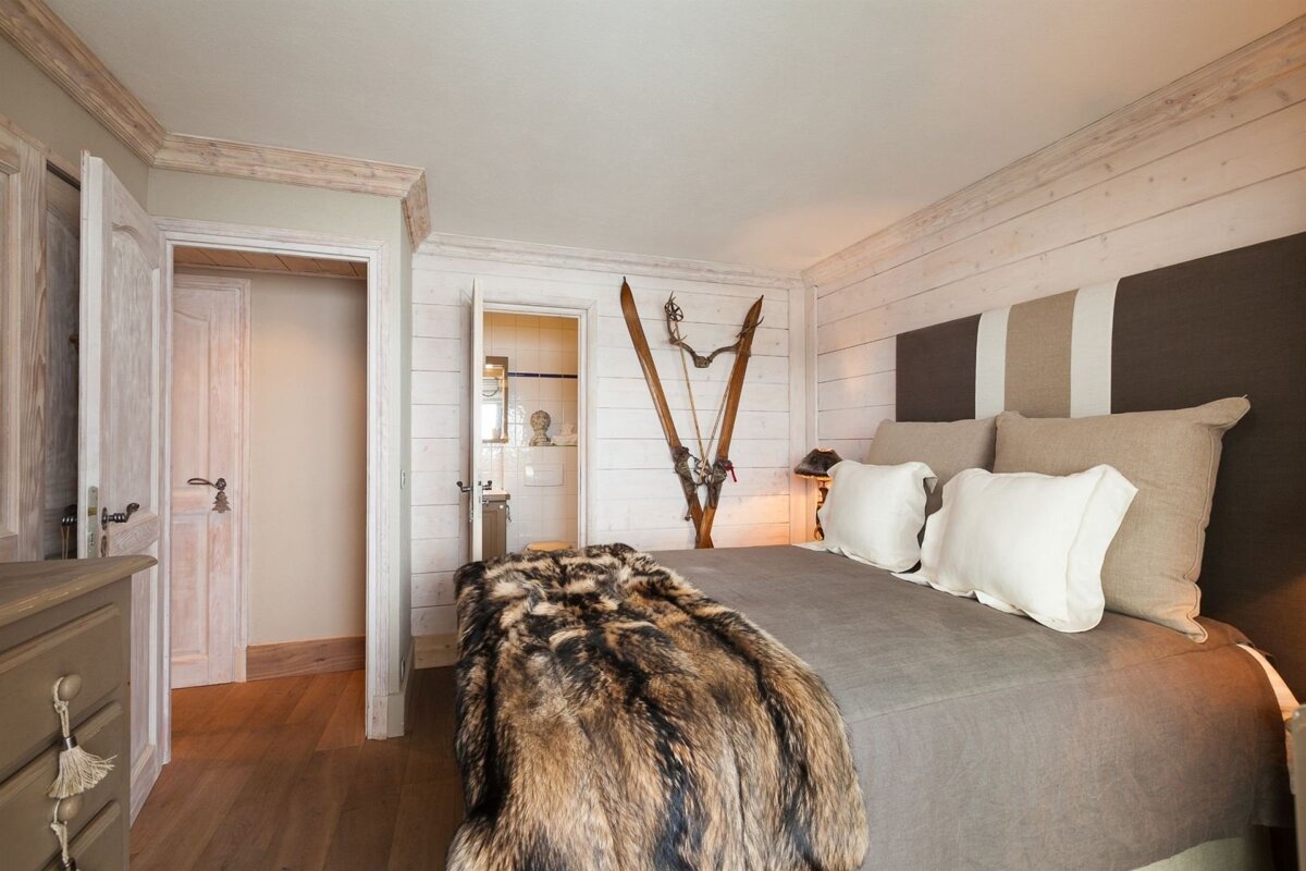 A rustic bedroom with a large bed, faux fur blanket, and antique skis on a white wood-paneled wall. Neutral tones create a cozy, lodge-like feel.