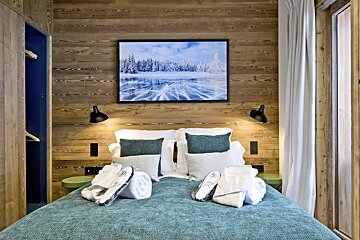 Cozy bedroom with wood-paneled walls, a teal bed topped with white towels, and a winter landscape picture hanging above.