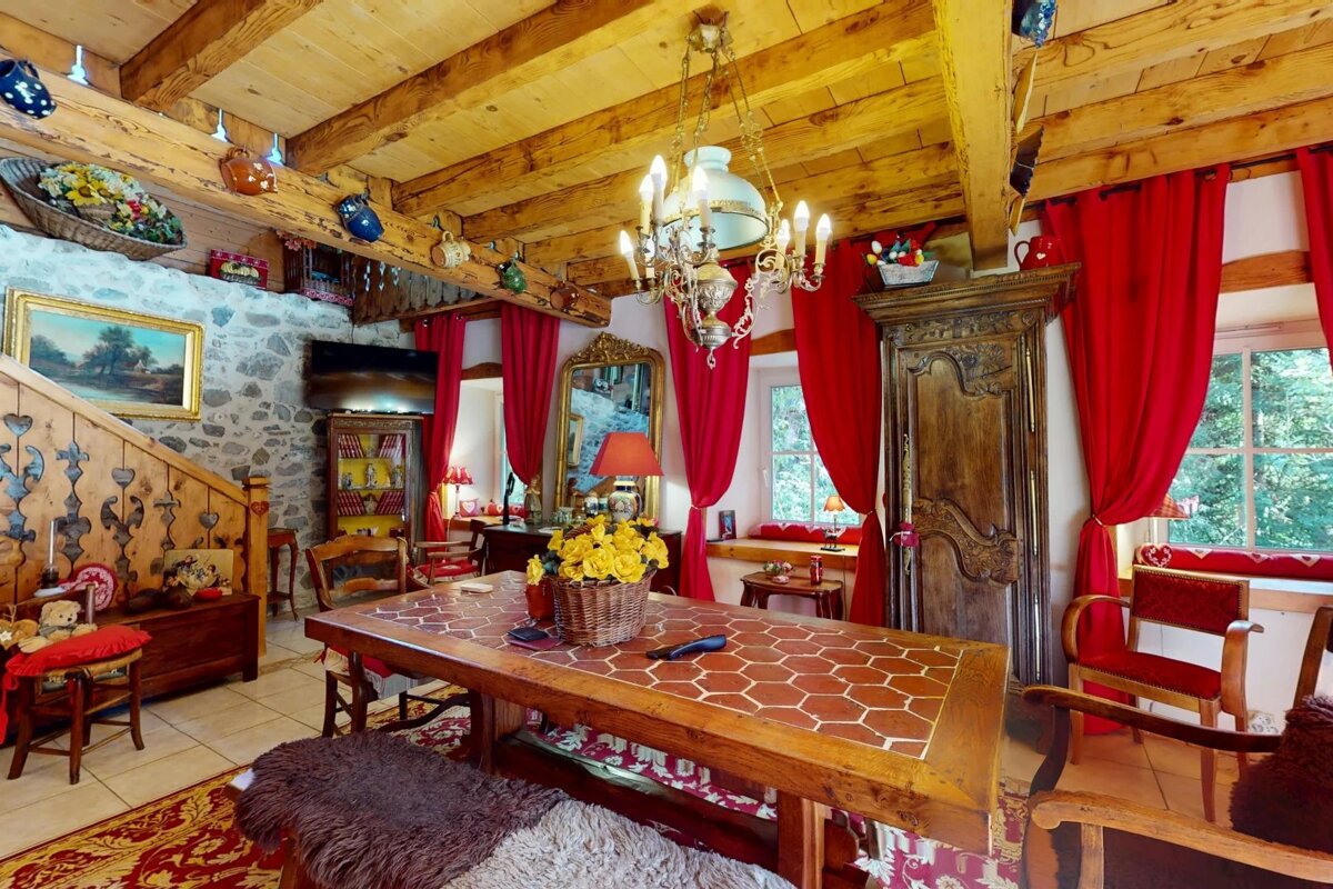 Cozy rustic dining room with wooden beam ceilings, bright red curtains, an ornate chandelier, and a tiled wooden table.
