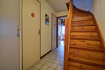 A narrow hallway with multiple doors, a tiled floor, and a wooden staircase on the right. One door has a red 