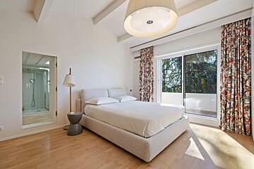 Bright, modern bedroom with a large bed, sliding door to a balcony, floral curtains, and a visible glass shower.
