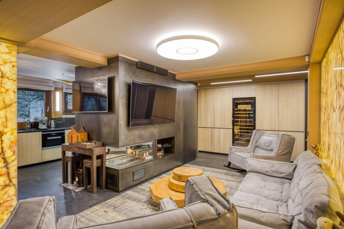 Luxurious open-plan living room with kitchen, a striking TV/fireplace unit, cozy gray seating, wood paneling, and an illuminated onyx wall.