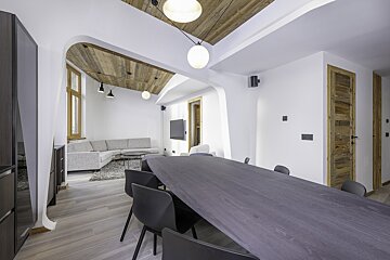 A modern, open-plan living and dining room features white walls, wooden ceiling accents, a long dark dining table, and a gray L-shaped sofa.