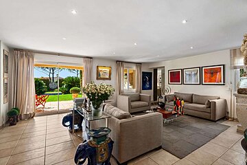 A modern living room with neutral sofas and colorful art, featuring large glass doors opening to a lush green garden and patio.