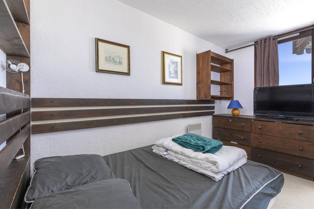 A cozy room features a single bed with gray bedding, white duvet, and teal blanket. White walls have dark wooden accents, framed art, and shelves. A dresser with a TV sits by a window revealing a blue