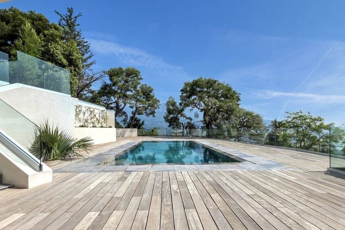 Luxurious rectangular pool set in a large wooden deck with glass railings, surrounded by green trees under a clear blue sky.