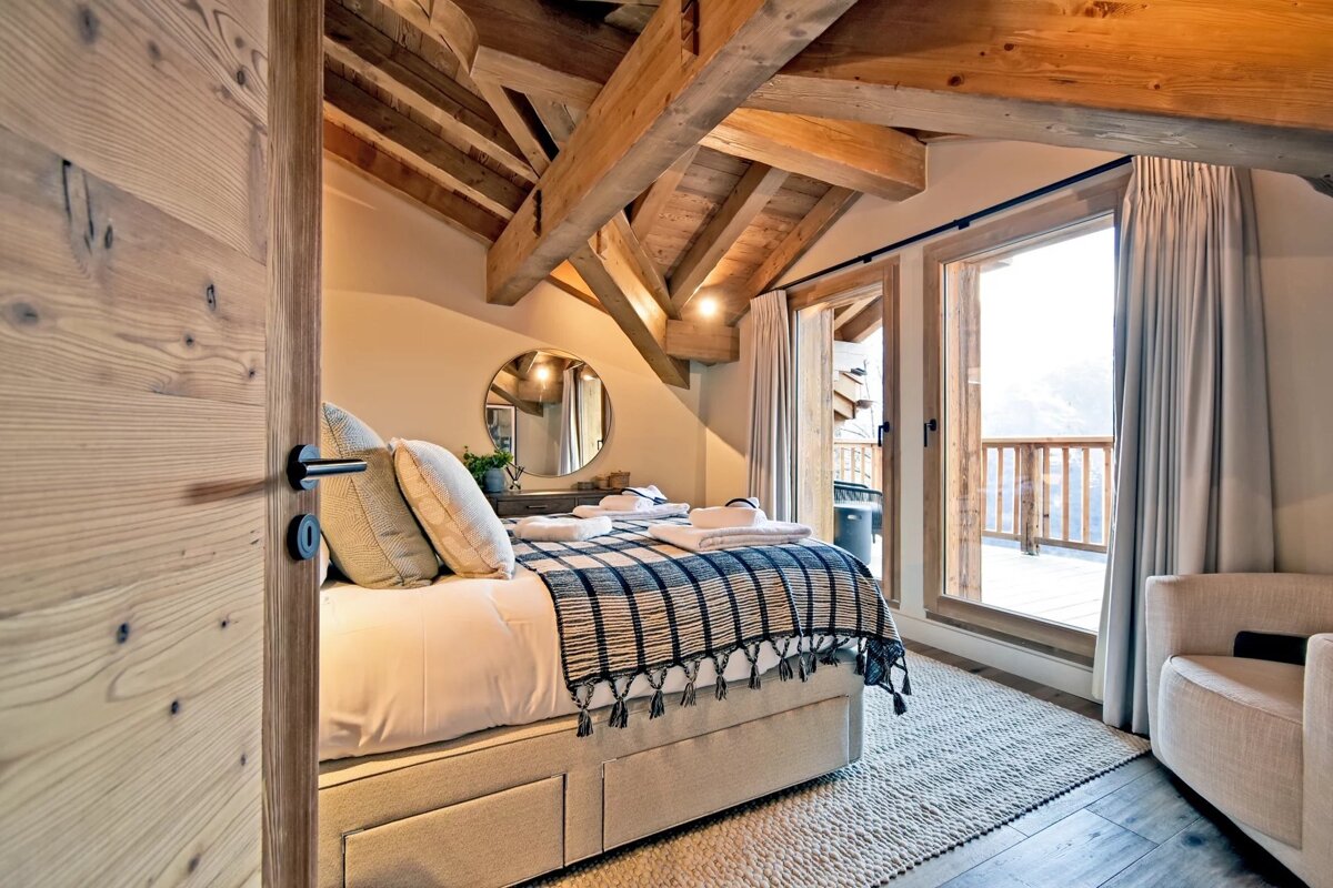 Cozy chalet-style bedroom with exposed wooden beams, a bed with a plaid throw, and glass doors opening onto a balcony.