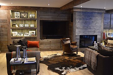 A cozy, rustic living room featuring reclaimed wood walls, a fireplace, TV, comfy sofas, leather chairs, and a cowhide rug, all warmly lit.
