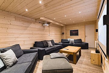 A cozy, wood-paneled media room features dark gray sofas, a wooden coffee table, projector, and screen, illuminated by recessed lighting.