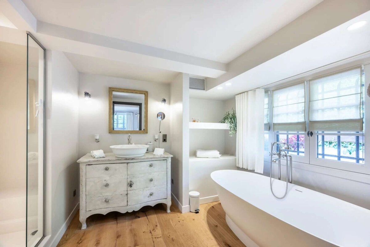 A chic, bright bathroom featuring a white freestanding tub, ornate vanity, wood floor, glass shower, and large window with blinds.