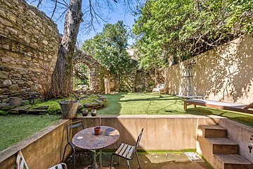 A rustic terraced garden features a sunken patio with a metal table and chairs, rising to a sunny artificial lawn bordered by old stone walls, an archway, and lush trees.