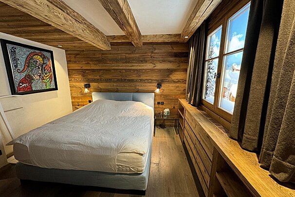 A rustic-chic chalet bedroom with wooden walls/beams, a large bed, modern art, and a window revealing a snowy mountain vista.