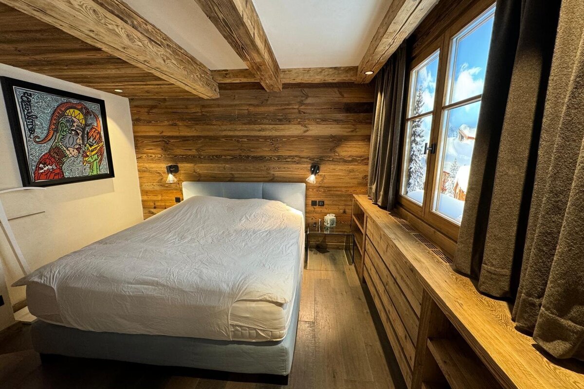 A rustic-chic chalet bedroom with wooden walls/beams, a large bed, modern art, and a window revealing a snowy mountain vista.