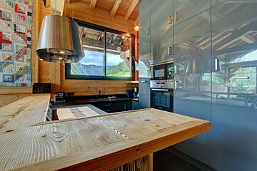A modern kitchen features glossy gray cabinets, built-in appliances, and a rustic wooden bar, walls, and ceiling, with a large window overlooking a lush mountain landscape.