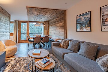Cozy alpine chalet living room with a grey sofa, nested wood coffee tables, wooden paneling, and a dining area overlooking a forest.
