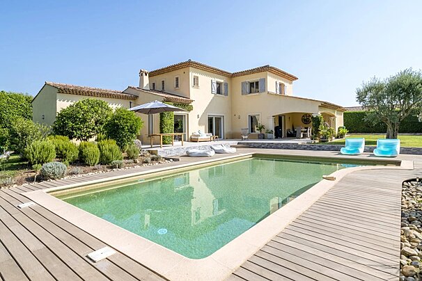 A sunny, light-colored house with a tiled roof, blue shutters, and a green swimming pool with a wooden deck. Lush garden and clear blue sky.