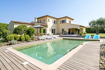 A sunny, light-colored house with a tiled roof, blue shutters, and a green swimming pool with a wooden deck. Lush garden and clear blue sky.