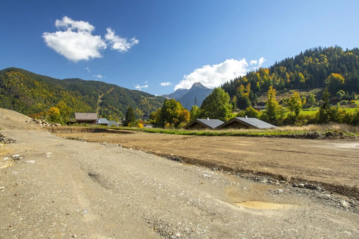 A cleared dirt plot in a scenic mountain valley, featuring wooden chalets, autumn forests, and a bright blue sky.