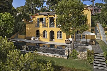 A vibrant yellow villa nestled amidst lush trees, featuring a pool, patio, and terraced gardens with stone walls and stairs.