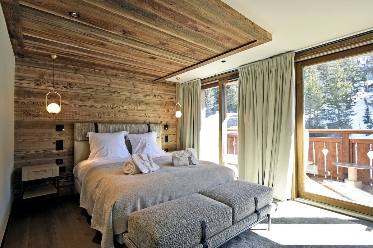 Cozy chalet bedroom featuring wood-paneled walls, a large bed, hanging lamps, and a glass door opening to a snowy balcony view.